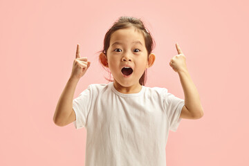 Sweet korean child girl looks amazed points at something up and looking to camera, isolated on a pink background. Cute little kid sees an amazing thing up, gestures indoor