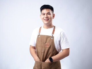 A young Asian man, dressed in a white shirt and light brown apron with leather straps, stands confidently with his arms crossed against a plain white background