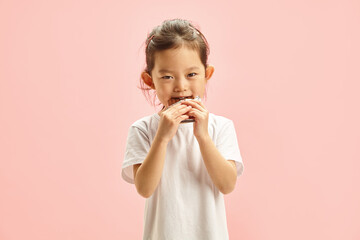 Photo of Cute Little Asian Girl Standing on a Pink Isolated Background, Eating a Chocolate Bar With an Appetite. Sweet Tooth Dilemma. Kids' Dental Health Alert.