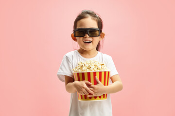 Joyful and Surprised Child Girl in 3D Glasses Holding Popcorn Bucket Standing Over Pink Isolated...