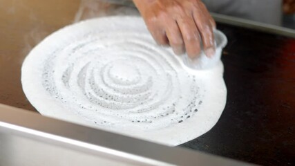 traditional South Indian dish, being cooked on a hot griddle. The dosa batter is being spread in a circular motion, with steam rising from the hot surface.