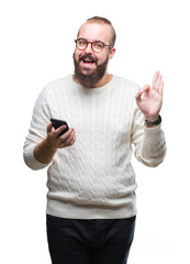 Young caucasian hipster man texting sending message using smartphone over isolated background doing ok sign with fingers, excellent symbol