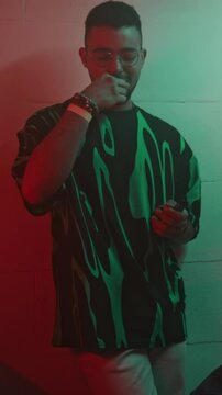 Vertical handheld lowkey shot of confident guy in glasses and vibrant t-shirt taking mdma pill and giving one to his girlfriend in trendy outfit while hanging out in corner of neon lit nightclub