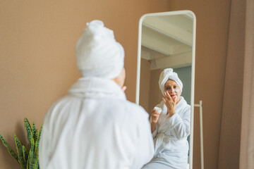 Mature adult old woman looking at mirror applying nourishing anti age face cream doing daily beauty...