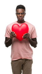 Young african american man holding red heart with a confident expression on smart face thinking serious