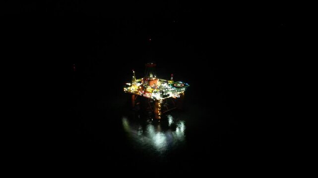 Large illuminated oil drilling rig Offshore facility for extraction of oil and gas night aerial footage downward panning far distance