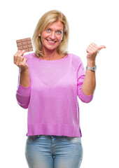 Obraz premium Middle age blonde woman eating chocolate bar over isolated background pointing and showing with thumb up to the side with happy face smiling