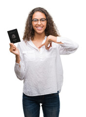 Young hispanic woman holding passport of United States of America with surprise face pointing finger to himself