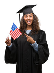 Young hispanic woman wearing graduation uniform holding flag of United States very happy pointing...