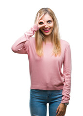 Young beautiful blonde woman wearing pink winter sweater over isolated background doing ok gesture with hand smiling, eye looking through fingers with happy face.