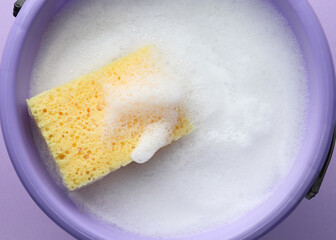 Bucket with foam and sponge on violet background, top view