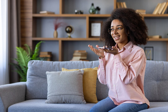 Woman sitting comfortably on couch using voice assistant on smartphone. Living room setting with bookshelf in background. Concept of technology, communication, modern lifestyle, and home comfort.