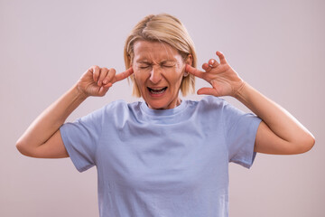 Portrait of frustrated mature woman covering her ears and shouting on a gray background.