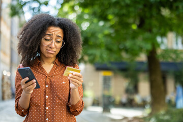 Upset young African American woman standing outside on the street, holding a cell phone and looking...