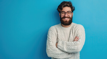 Obraz premium Bearded young man in a casual sweater and glasses, arms crossed, smiling warmly, looking at the camera on a blue background