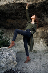 empowered woman in green coat with arms outstretched standing on rocky peak under blue sky