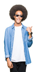 Young african american man with afro hair wearing thug life glasses smiling with happy face looking and pointing to the side with thumb up.