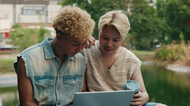 Hapy Asian LGBTQ friends using a laptop and enjoying coffee together in a park. Celebrating pride and connectivity