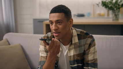 Happy African American man at home couch sofa biracial male ethnic guy using cellphone answering on audio message record voice mail recording answer in social media voicemail use speakerphone aloud