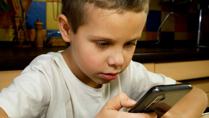 A boy in the kitchen with a phone in his hands taps the screen and talks. Communication using gadgets and Internet applications. Children and games in mobile phones.