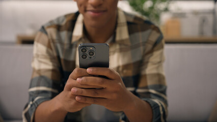 Happy smiling positive African American man at apartment couch smile biracial male ethnic guy user at home sofa using mobile phone looking at smartphone screen think ponder shopping online choose app