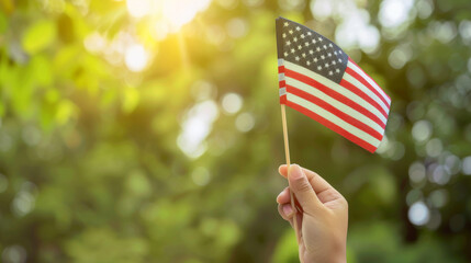 A hand proudly waving a small American flag against a sunlit, leafy background, symbolizing patriotism and national pride.