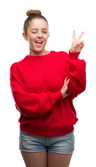 Young blonde woman wearing bun and red sweater smiling with happy face winking at the camera doing victory sign. Number two.