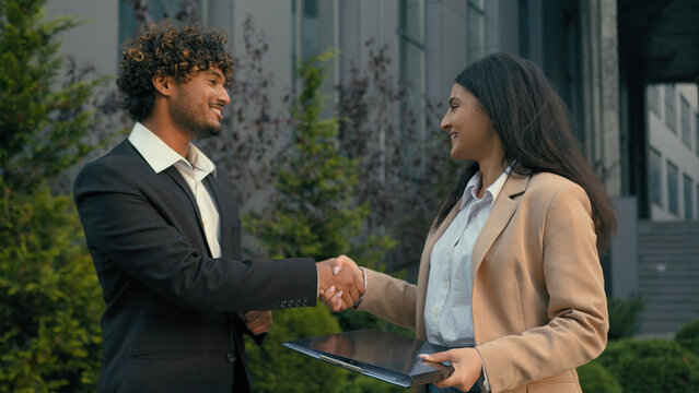 Two business people in city Arabian woman businesswoman intern holding folder papers documents talking with Indian businessman man leader investor talk shaking hands smiling handshaking contract agree