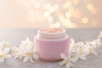 Cream in jar and beautiful jasmine flowers on grey table against light background with blurred lights, closeup