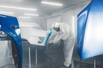 Car paint worker spraying blue paint to car body element using spray gun in vehicle workshop...