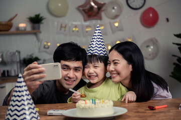 Happy Asian Family celebrating a birthday together at home. Young Asian father using smartphone taking selfies with son and wife while celebration birthday party of his son at home, social isolation.