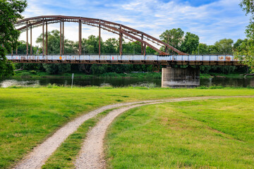 Fototapeta premium A dirt road running through a green meadow against the backdrop of a river and a railway bridge. Blue sky with white clouds..