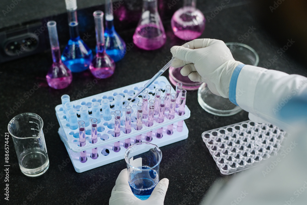 Aufkleber High angle of gloved hand of scientist with pipette dropping liquid into flask with chemical substance while standing by workplace