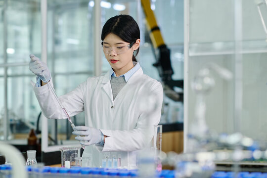 Young serious researcher in protective eyewear looking at liquid substance in flask while carrying out scientific investigation in laboratory