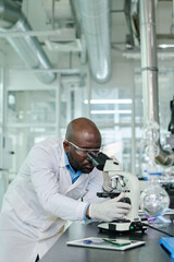Young serious male researcher in lab coat standing by workplace with microscope and looking at bacteria or new covid strain