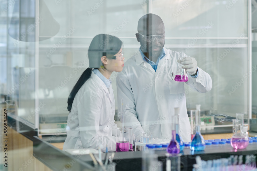 Poster young intercultural biochemists in lab coats looking at beaker with liquid substance while studying 