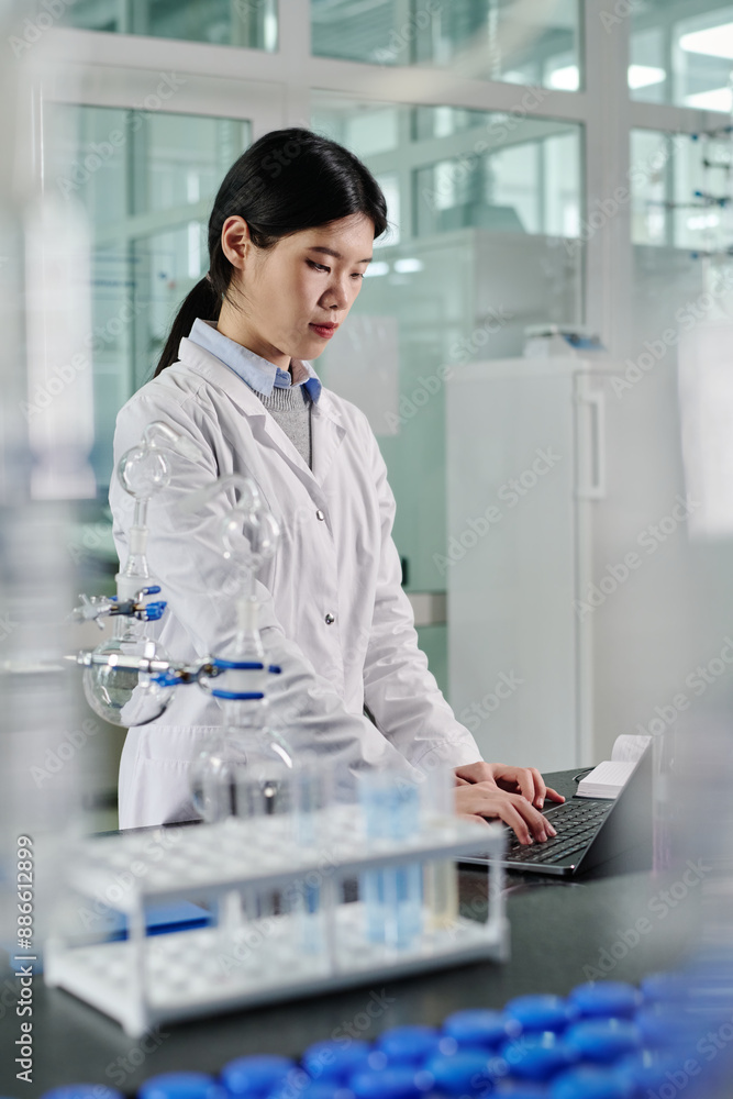 Poster Young female laboratory worker standing by workplace and looking at laptop screen while entering results of experiment in database