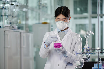 Young Chinese woman in lab coat, protective gloves and respirator studying characteristics of...
