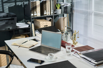 Laptop with blank screen standing on desk with number of other office supplies, documents, open notebook, statuette of Justice goddess