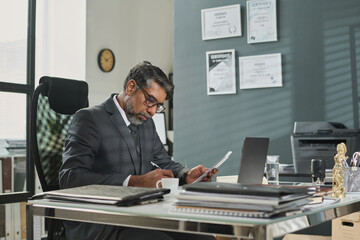 Mature man in suit and eyeglasses sitting by workplace in office and signing juridical paper document after reading it