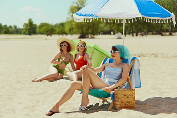 Three women in retro swimwear enjoying sunny beach day. One sits under umbrella with drink, while...