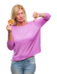 Middle age blonde woman eating sweet waffle over isolated background with angry face, negative sign showing dislike with thumbs down, rejection concept