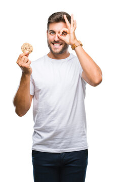 Young handsome man eating chocolate chips cookie over isolated background with happy face smiling doing ok sign with hand on eye looking through fingers