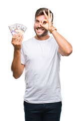Young handsome man holding stack of dollars over isolated background with happy face smiling doing ok sign with hand on eye looking through fingers