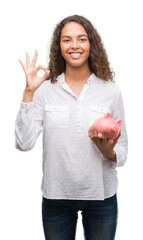 Young hispanic woman holding piggy bank doing ok sign with fingers, excellent symbol