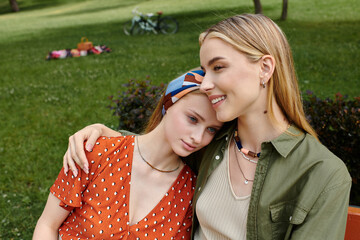 A youthful lesbian couple cuddles in a lush park, one woman in a red polka-dot dress resting her head on her partners shoulder