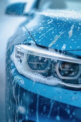 Blue car exterior close-up with soapy water covering headlights and taillights. Glossy appearance reflects light in outdoor setting with blurred trees and buildings visible through windshield.