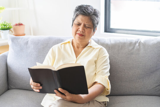 Presbyopia, Hyperopia mature, aged asian woman having vision problem trying to stare read text on a book on sofa at home, eye disease of elderly. Poor eyesight farsightedness,  health care concept