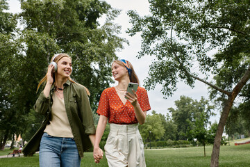 A young lesbian couple enjoys a carefree stroll through a lush green park, hand in hand, as they listen to music and share a laugh.