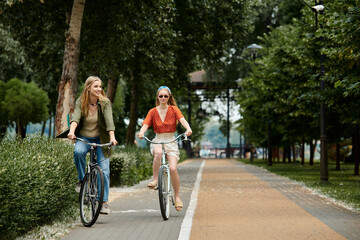 Obraz premium Two young women ride bicycles side-by-side on a paved path through a lush green park.